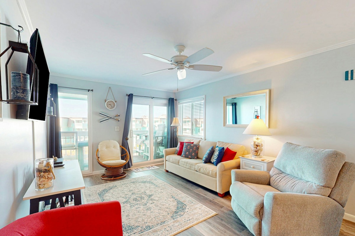 A spacious living room features comfortable seating options, including a light-colored couch and a plush armchair. A large TV is mounted on the wall opposite a decorative rug. Natural light floods the area through large windows, enhancing the bright atmosphere.