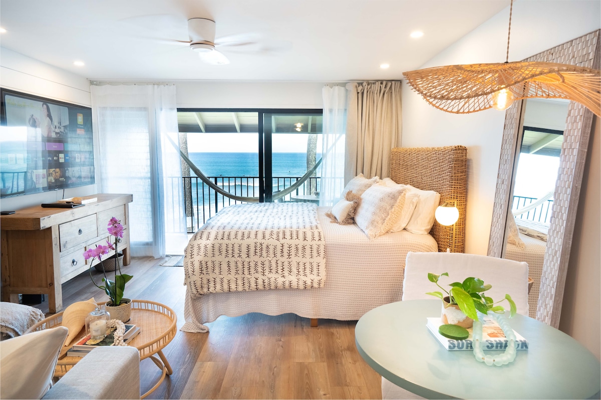 A bright studio space features a spacious king-sized bed with coastal-themed bedding. Large windows offer unobstructed ocean views, while a hammock hangs on the lanai. A small dining table with greenery and a light wood console completes the inviting ambiance, enhancing the serene coastal setting.