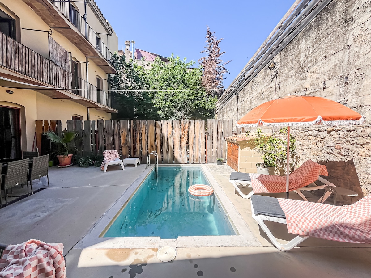 An outdoor pool area is framed by stone walls, with a refreshing blue pool at the center. Sun loungers with striped cushions are situated nearby, and a bright orange umbrella provides shade. Lush greenery is visible in the background.