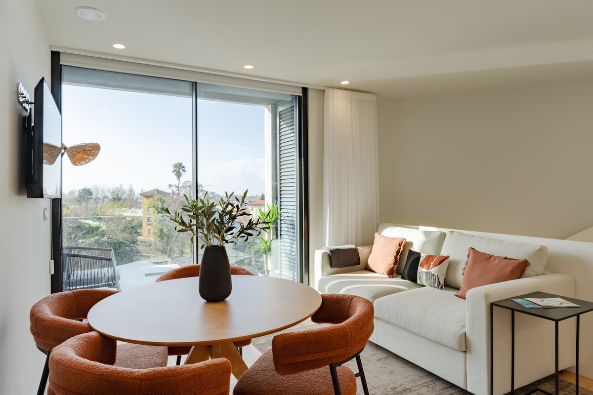 A living area featuring a round dining table surrounded by four orange chairs. A light-colored sofa with decorative cushions is positioned nearby, along with a side table. Large glass doors provide views of the outdoors, enhancing the space with natural light.