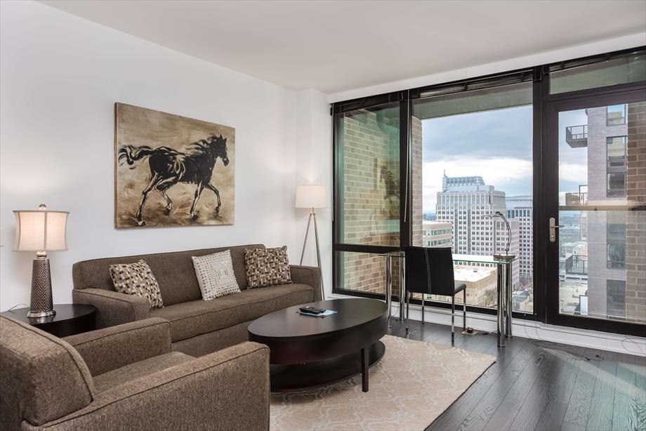 A modern living area is presented with a comfortable sofa and a round coffee table. A large window offers a view of the surrounding city, while a framed artwork of a horse adorns the wall. Warm lighting is provided by a stylish lamp.