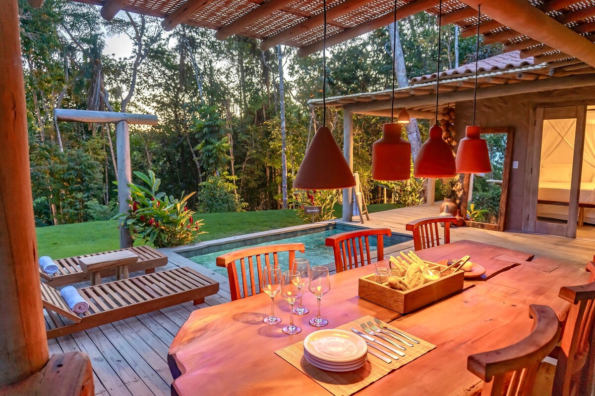 An outdoor dining area with a wooden table is illuminated by red pendant lights. The setting overlooks a lush green landscape and a swimming pool. Surrounding the space are wooden decks and tropical plants, creating a serene atmosphere.