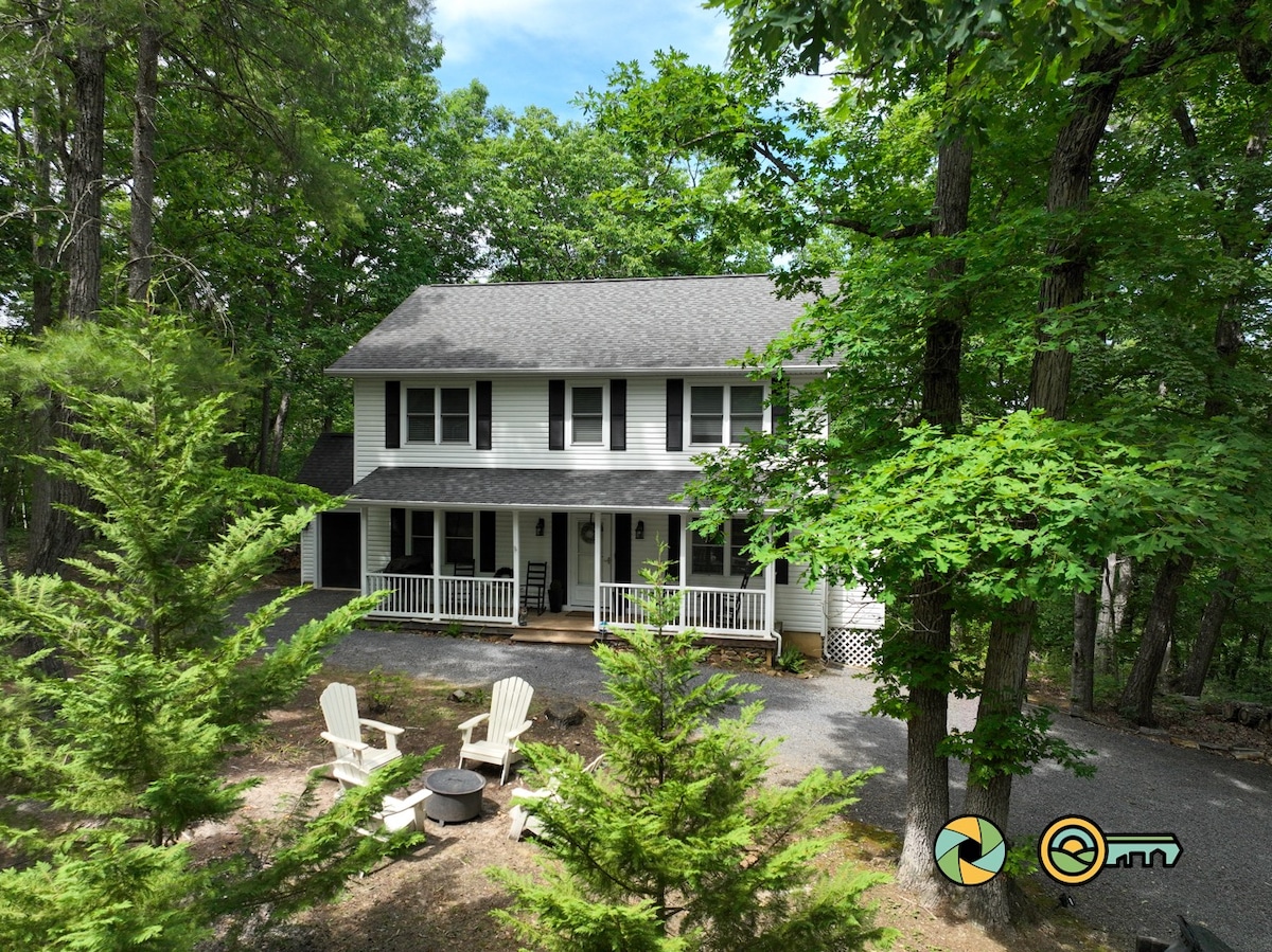 The exterior of a two-story home is surrounded by lush trees, providing a sense of privacy. The covered porch features adirondack chairs, and a fire pit is situated in the front yard. The driveway is framed by greenery, creating a welcoming entrance.
