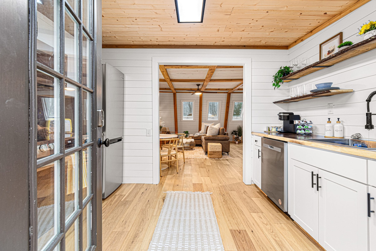 An inviting entryway features light wood flooring leading towards a bright, open kitchen. White cabinetry is complemented by a wood countertop and stainless-steel appliances. A cozy living area is visible in the background, accented by natural light streaming through large windows.
