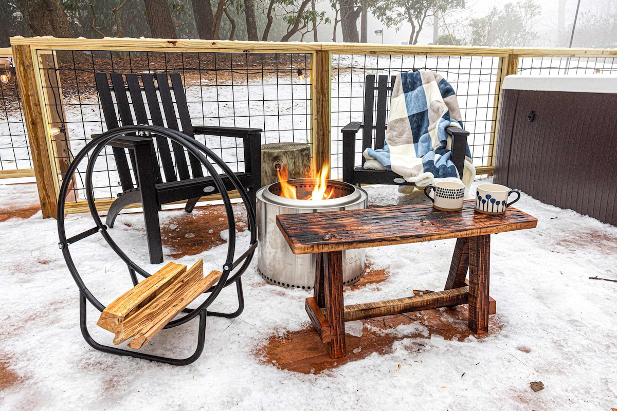 An outdoor seating area features a wooden bench positioned next to a circular fire pit with flickering flames. The space is adorned with black rocking chairs and a cozy plaid blanket, set against a snowy background.