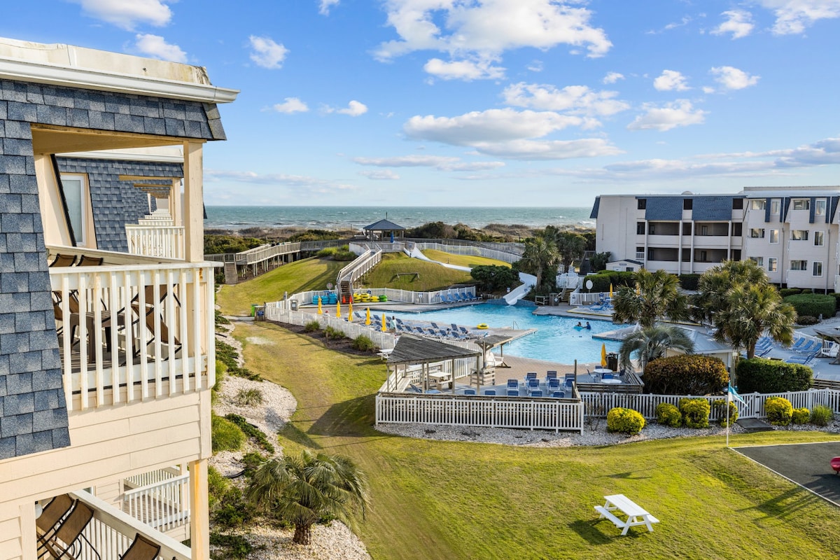 A scenic view of the pool area and expansive lawns is presented from the balcony. The inviting pool is surrounded by lounge chairs and colorful umbrellas, with the Atlantic Ocean visible in the background under a partly cloudy sky.