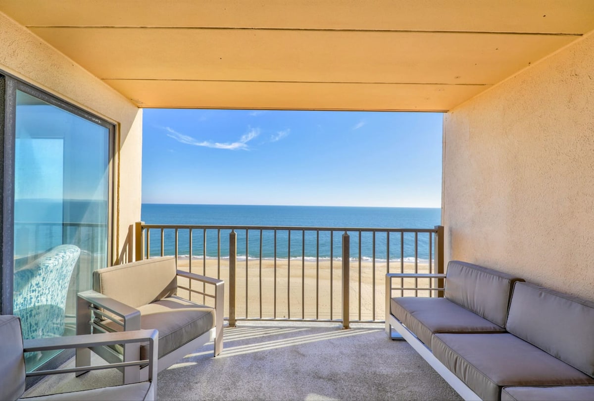 A private balcony is showcased with modern outdoor lounge furniture, providing seating for five. The expansive view reveals a vast ocean meeting a sandy beach, framed by clear blue skies, creating a serene outdoor space for relaxation.