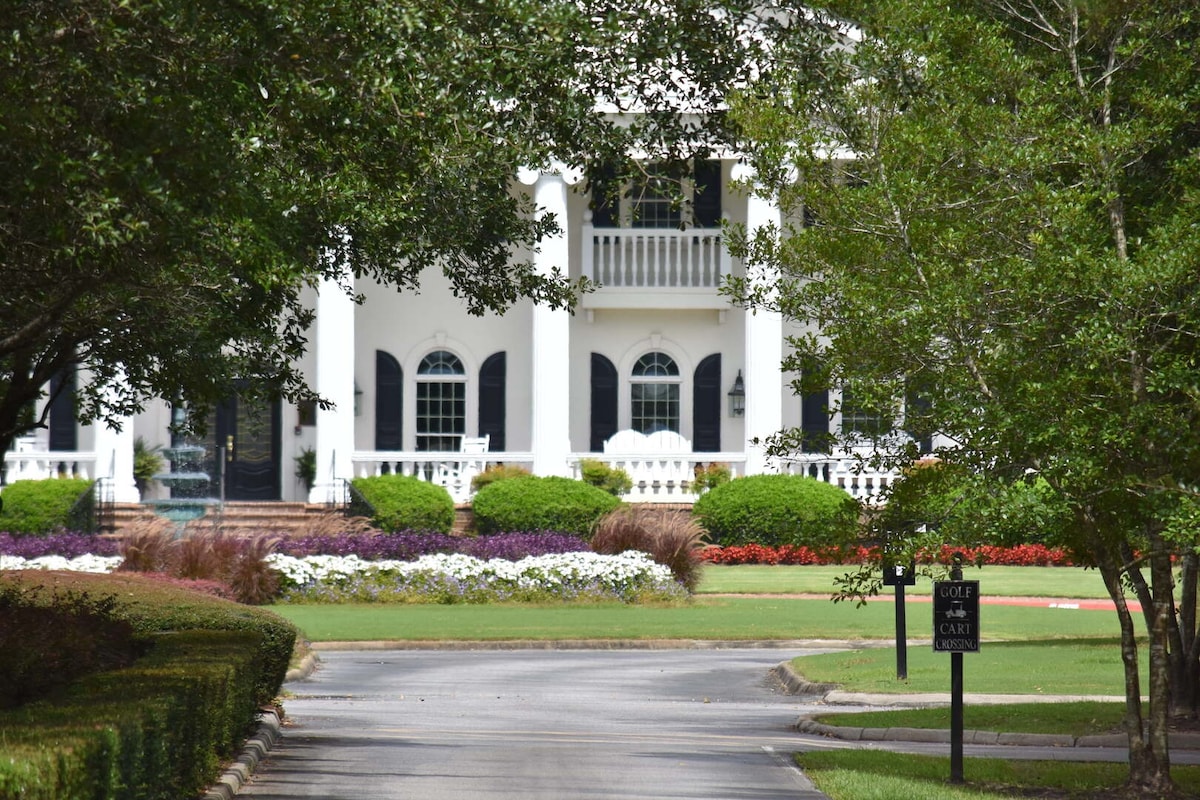 A well-maintained driveway leads toward a grand white building, framed by lush greenery and colorful flowerbeds. Black shutters contrast with the white facade, and a wide staircase provides access to the entryway. A sign indicating 'Guest Check In' is visible along the path.