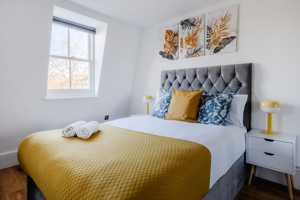 A cozy sleeping area features a spacious bed draped in a yellow quilted coverlet. Decorative pillows in blue and white are arranged on the bed. Two bedside lamps stand on either side of the bed, and wall art with a botanical theme complements the decor.