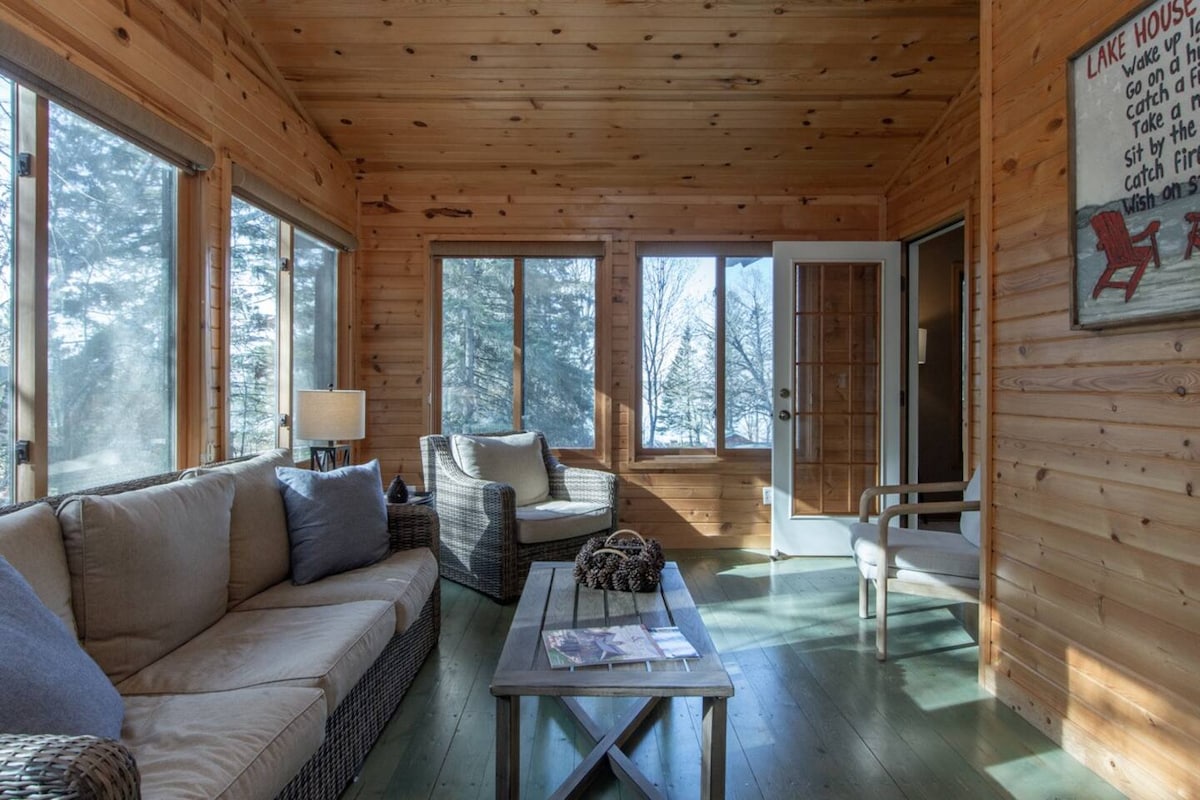 A sunlit living area features wooden walls and large windows that invite natural light. Two comfortable seating options, a sofa and an armchair, are arranged around a low table displaying magazines. A lamp provides additional lighting, enhancing the cozy cabin atmosphere.