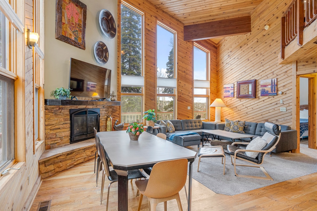 The open-concept living area features a stone fireplace and large windows that allow natural light to fill the space. A comfortable sectional couch and an elegant dining table are surrounded by wood accent walls, creating a warm atmosphere for relaxation and gatherings.