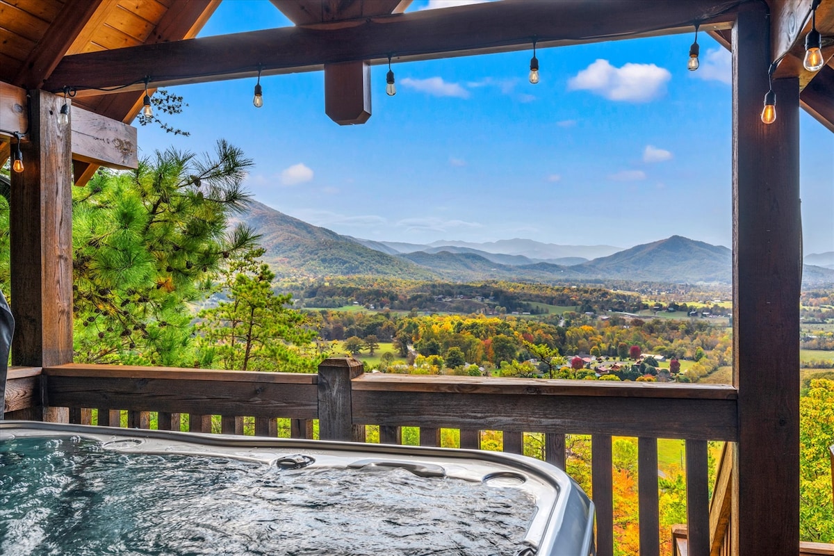 A hot tub is positioned on a wooden terrace, overlooking a picturesque view of rolling mountains and vibrant autumn foliage. The scene is enhanced by strings of lights hanging above, contributing to a tranquil outdoor space ideal for relaxation.
