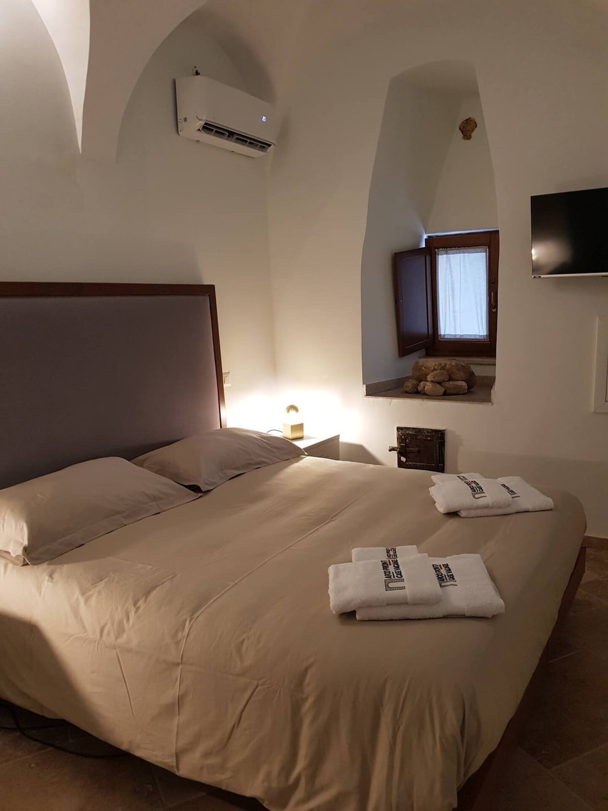 A spacious sleeping area is defined by a large bed with neutral-toned linens. Folded towels are positioned neatly at the foot of the bed. A small window allows natural light to enter, complemented by a flat-screen TV mounted on the wall.