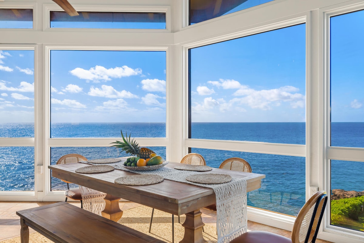 A dining area features a wooden table surrounded by rattan chairs, showcasing a simple table setting. Expansive windows frame stunning ocean views, allowing natural light to fill the space. The blue sea and sky, along with scattered clouds, create a serene backdrop.