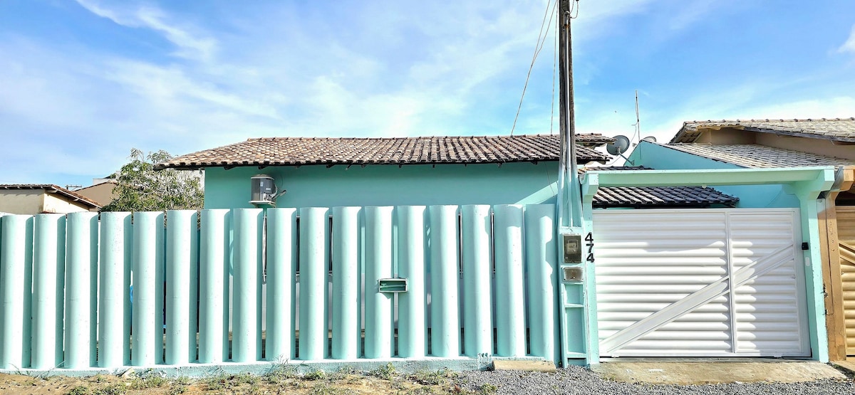 A light blue exterior of the house is featured with a distinctive white fence that has vertical slats. A private garage door is visible, alongside a mailbox on the fence. Clear skies can be seen above, enhancing the tranquil neighborhood setting.