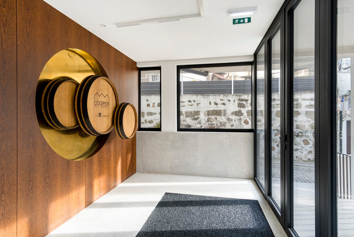 A modern entryway features a large wooden wall display with circular wine barrels. Floor-to-ceiling windows allow natural light to illuminate the polished floor and create an open, airy atmosphere, while a textured mat welcomes visitors at the entrance.