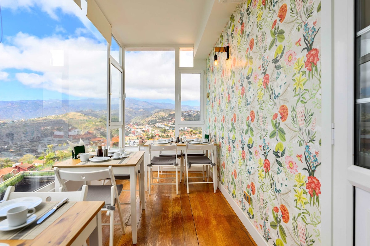A bright dining area features large glass windows offering expansive views of the surrounding mountains and town. Floral wallpaper adds a touch of color to the space, which includes wooden tables and chairs arranged for dining. Natural light fills the room, creating a welcoming ambiance.