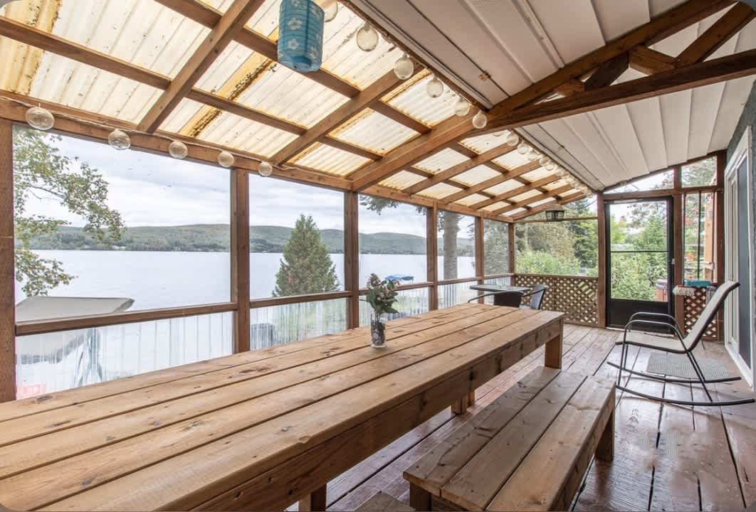A spacious covered porch features wooden picnic-style tables and benches, framed by large windows that offer views of the lake. Soft, natural lighting flows in from above, creating an inviting space for gatherings. Decorative string lights and a simple vase with flowers add a touch of charm.