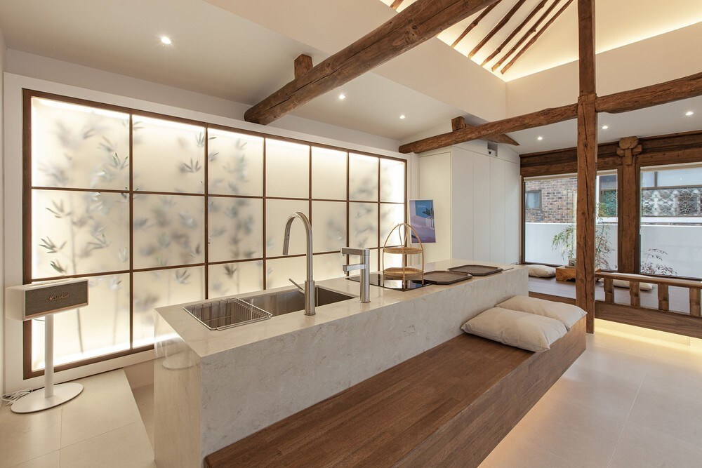 The kitchen features a central island made of stone with dual sinks and minimalistic fixtures. A wooden bench runs alongside the island, and ambient lighting highlights the modern cabinetry. Frosted glass panels with a nature-inspired design enhance the space's aesthetic while allowing natural light to filter through.
