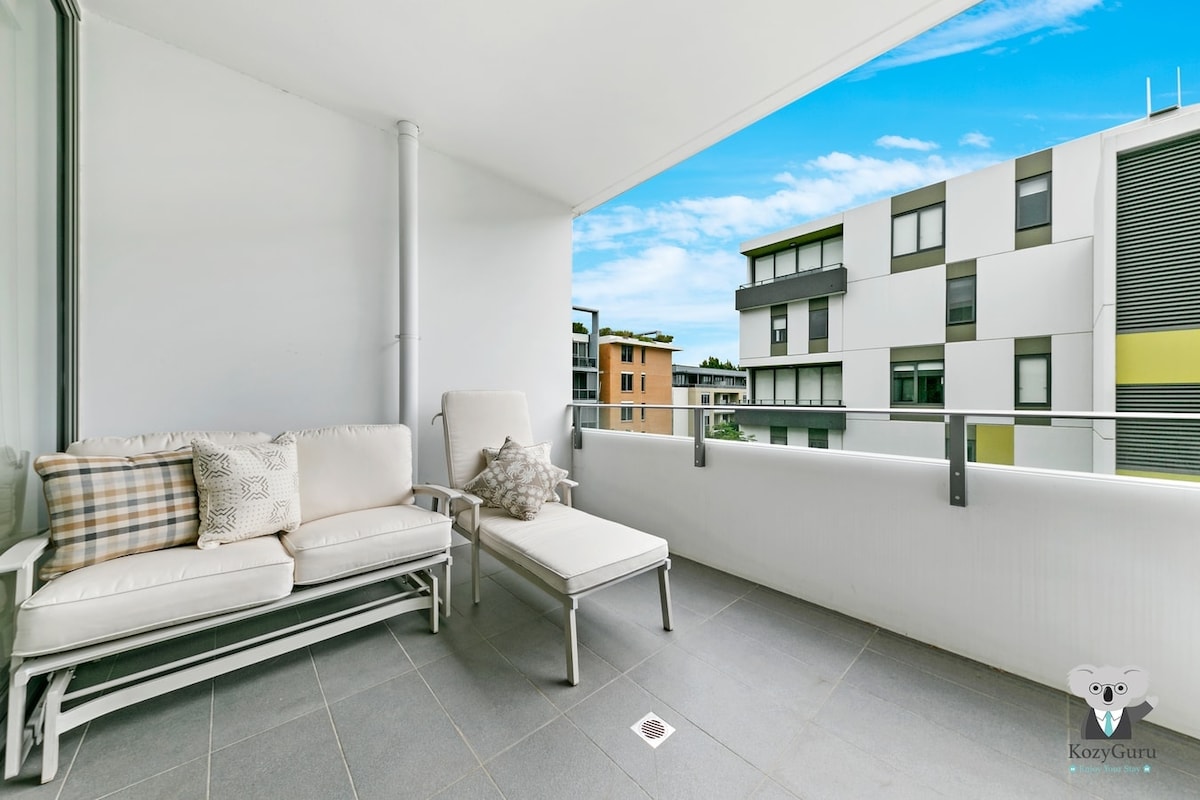A private balcony space features a light-colored sofa and a lounge chair, both positioned next to a railing. The tiled floor provides a modern touch, while a clear view of surrounding buildings and an open sky enhances the area’s ambiance.