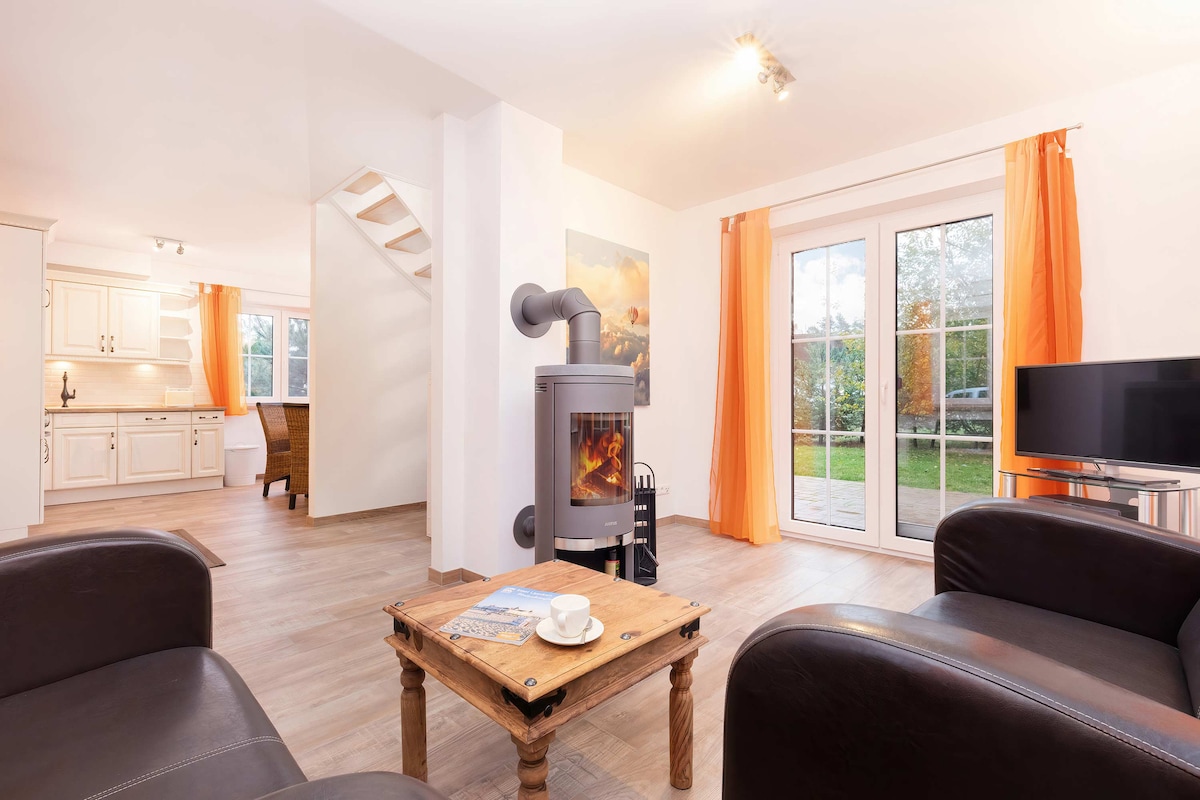 A bright living area features a stylish wood-burning stove at the center, surrounded by comfortable black leather chairs. Large glass doors lead to the outdoor space, while warm orange curtains add a touch of color to the neutral walls. A wooden coffee table is placed in front.