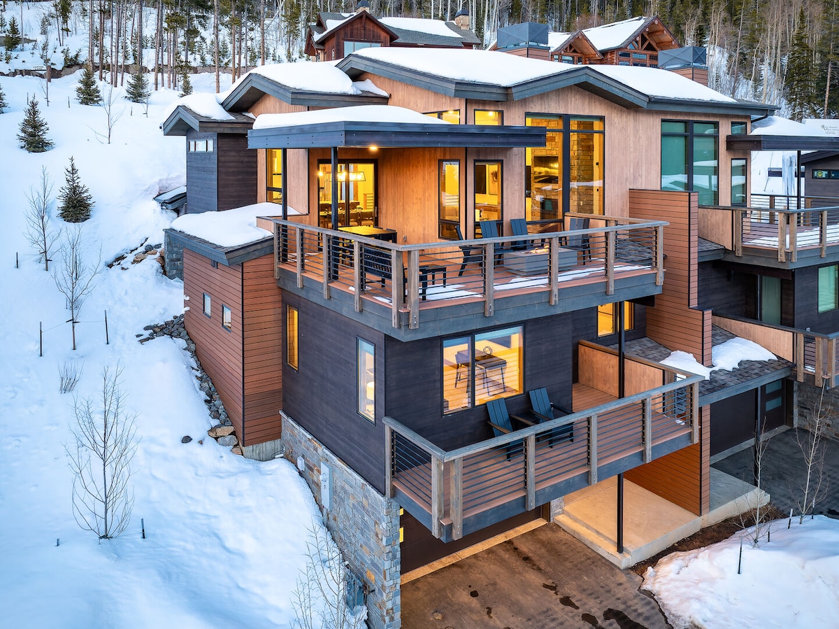 An exterior view of the mountain townhome showcases sleek modern architecture set against a snowy landscape. Multiple balconies extend from the property, offering seating areas with stunning views. Large windows reflect the surrounding winter scenery, while warm light emanates from inside, creating an inviting atmosphere.