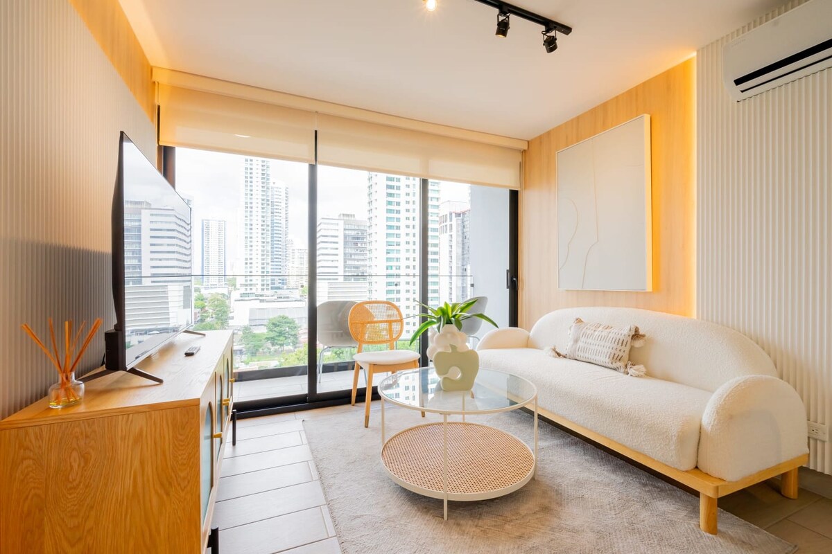 The living room is characterized by a large window offering city views, complemented by a comfortable, plush sofa and a decorative coffee table. A light wood cabinet spans one wall, while a flat-screen TV is mounted above. Neutral tones create a serene ambiance.