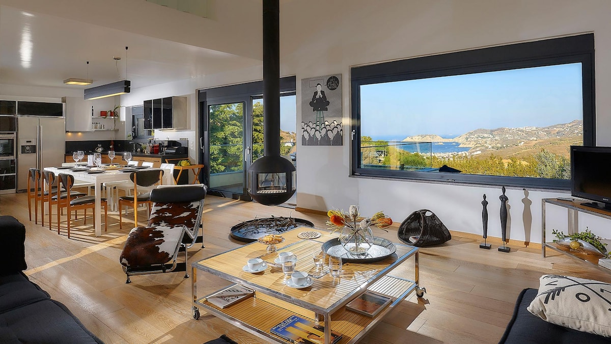 A spacious living area is presented, featuring large windows that offer scenic views of the sea. A central fireplace is surrounded by a stylish coffee table and comfortable seating. A dining table accommodates several chairs, while the kitchen area is visible in the background.