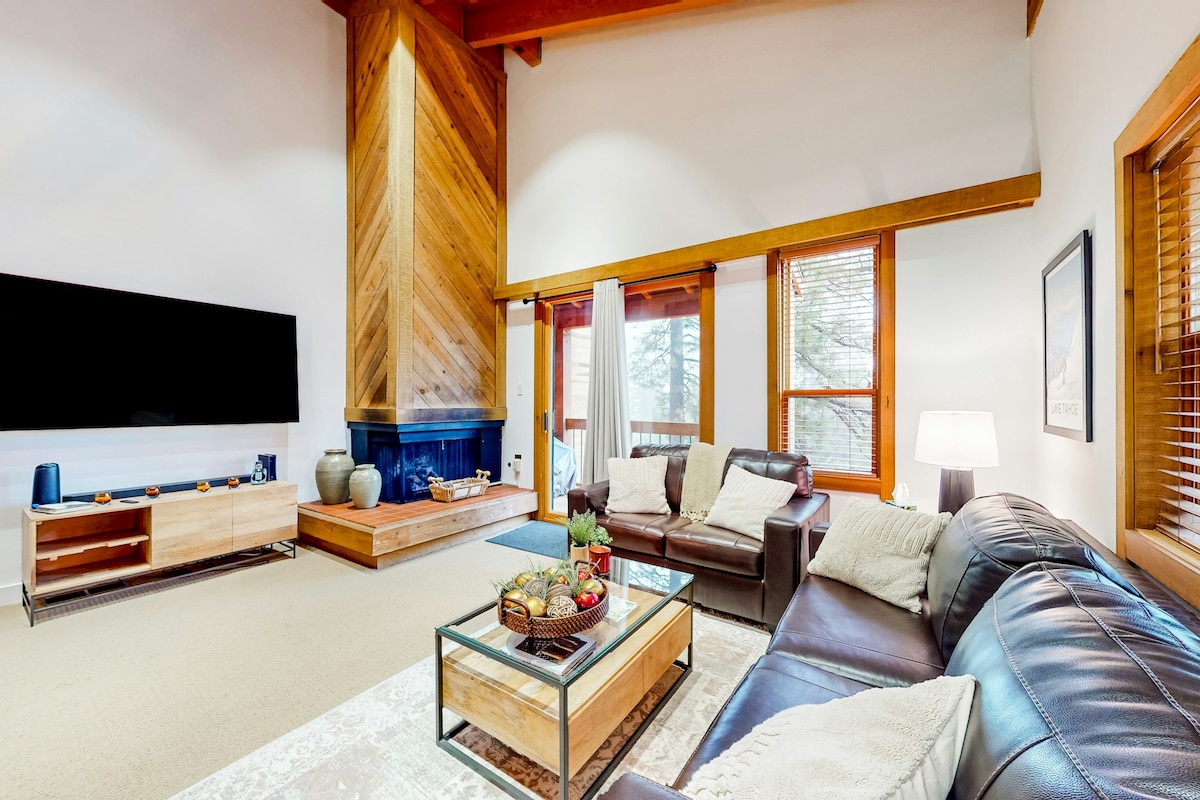 The living area features high ceilings and large windows, allowing natural light to fill the space. A modern leather sofa and a comfortable chair surround a glass coffee table, which holds a decorative fruit arrangement. A flat-screen TV is mounted on the wall, with a wooden console beneath it. A cozy fireplace is integrated into the wall, contributing to the inviting ambiance.