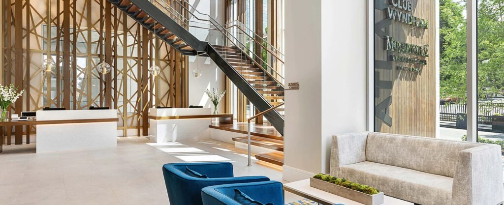 The lobby features a modern design with high ceilings and large windows, allowing natural light to fill the space. A sleek reception desk is paired with a comfortable seating area that includes a light-colored sofa and two blue chairs. A decorative plant adds a touch of greenery.