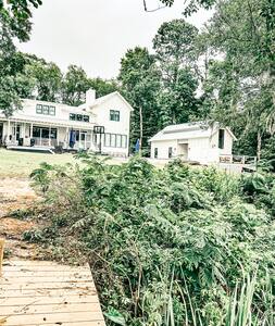 A serene view of a white two-story house and a separate building set against a lush green backdrop. The property features a wooden dock leading to a waterway, surrounded by diverse foliage and trees, creating a tranquil natural setting.