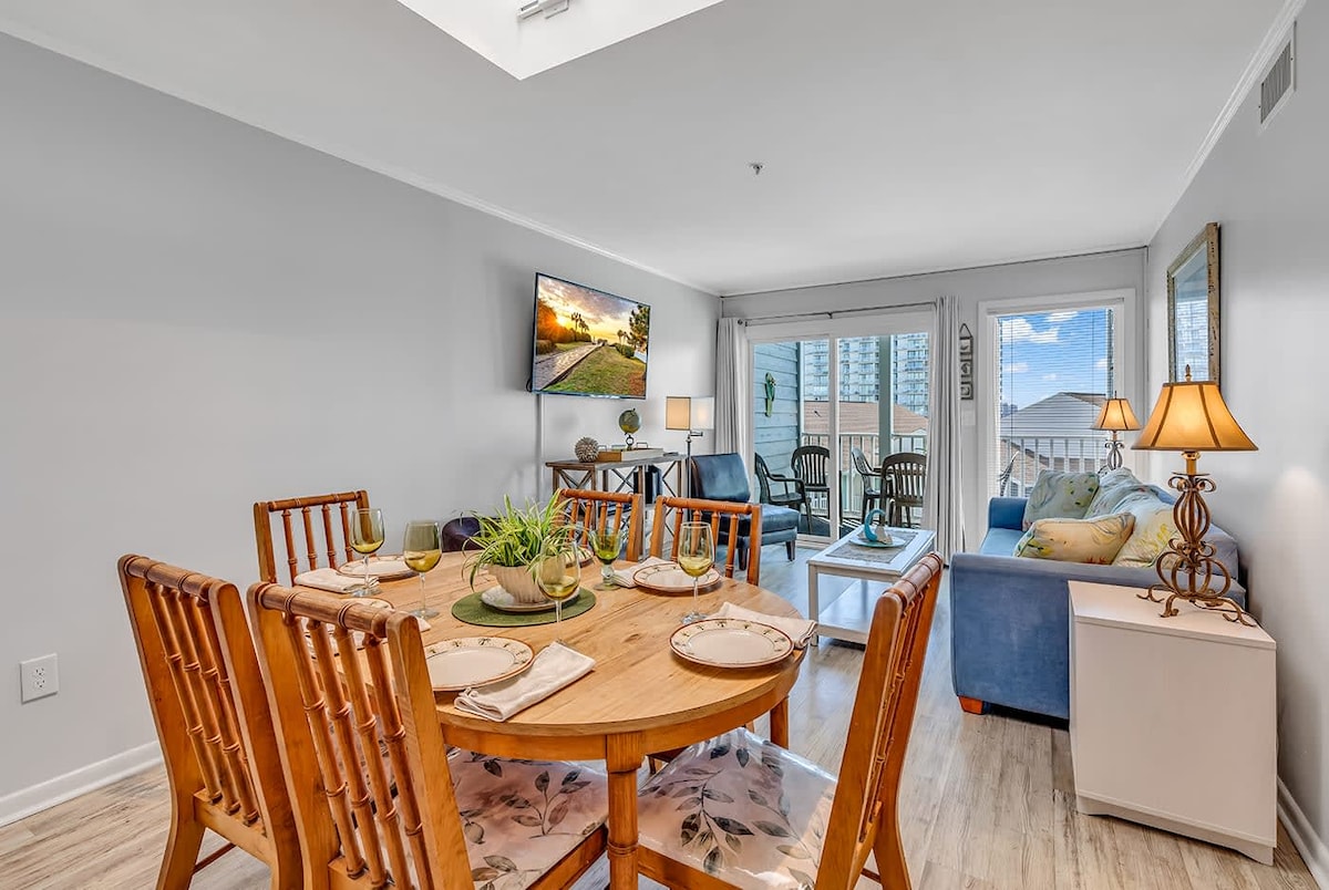 A dining area features a round wooden table set for four, surrounded by wooden chairs. A television is mounted on the wall, and natural light enters through sliding glass doors that lead to a balcony. A comfortable seating area with a blue sofa is also visible.