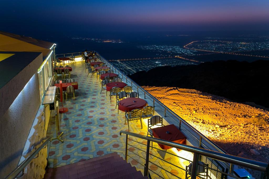 An outdoor terrace features several tables surrounded by scenic views of the city below. The space is illuminated by soft lights, with tiled flooring adding to the ambiance. The horizon showcases a gradient of colors as day transitions to night.