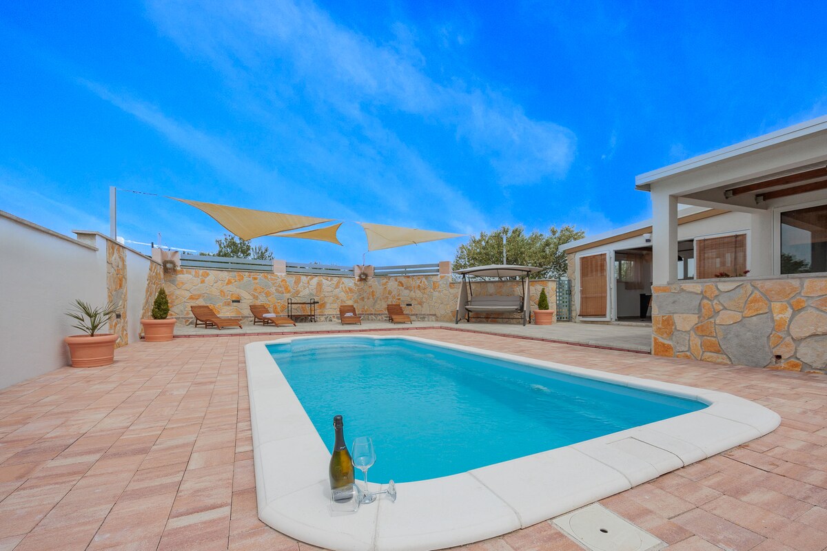 A private pool, surrounded by a stone-paved area, offers a place for relaxation and refreshment. Shade is provided by fabric canopies above. Potted plants are positioned around the pool, and a seating area is visible in the background, enhancing the outdoor space.