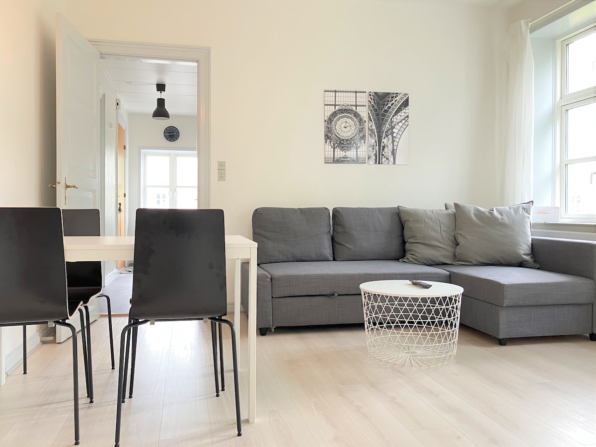 A modern living area is shown, featuring a gray sectional sofa adorned with cushions. A small round coffee table sits at its center, and a white dining table with four black chairs is positioned nearby. Light streams in through large windows, illuminating the space.