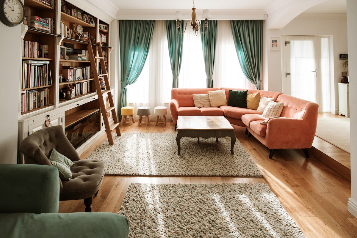A spacious living area features a coral-colored sectional sofa adorned with decorative cushions. A wooden table sits centrally on a textured area rug. Natural light streams through large windows, framed by soft, emerald drapes. A well-stocked bookshelf and a ladder add character to the room.