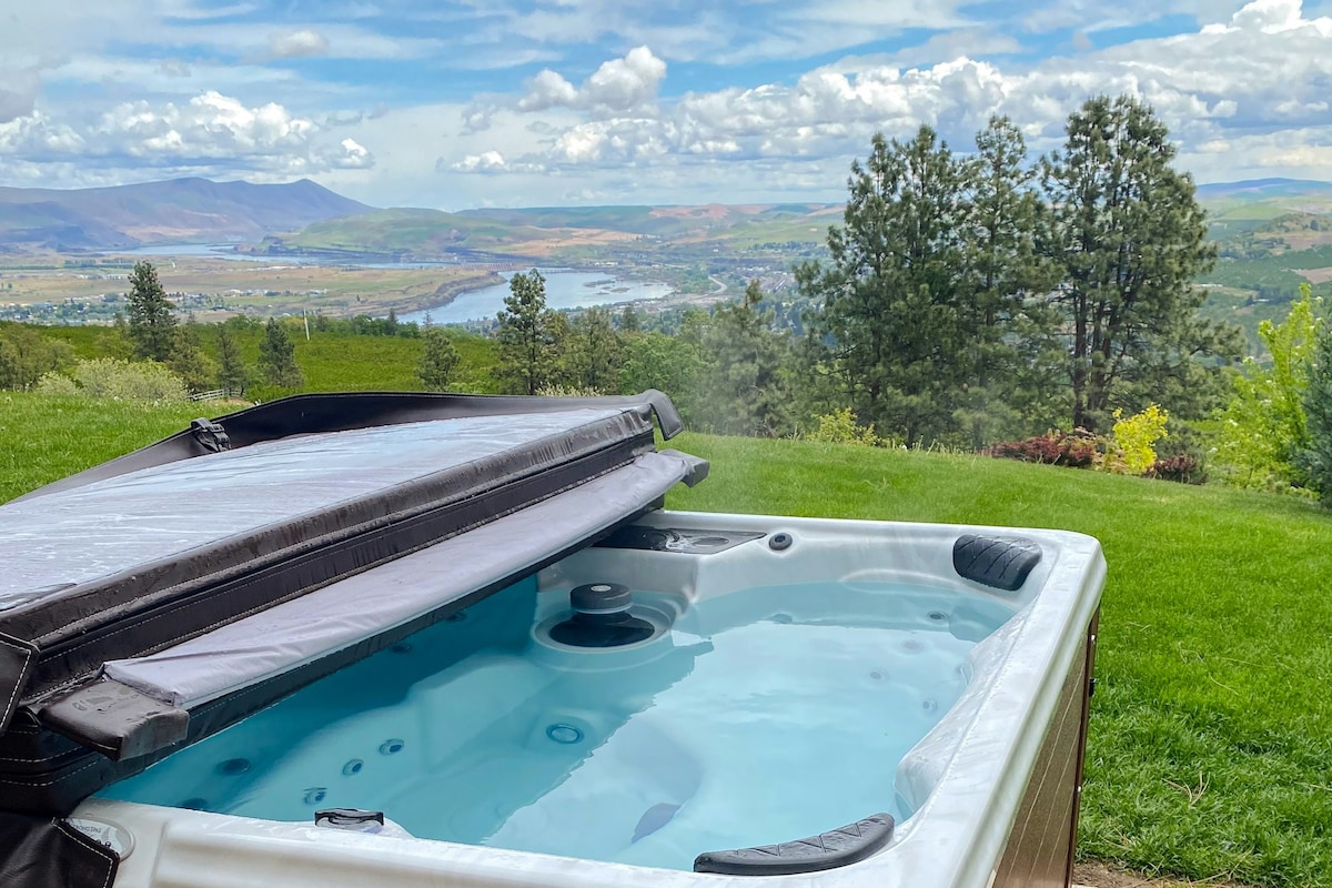 A hot tub is featured in the foreground, with steam rising gently from its surface. In the background, rolling green hills and the Columbia River are visible, framed by trees against a cloudy sky.