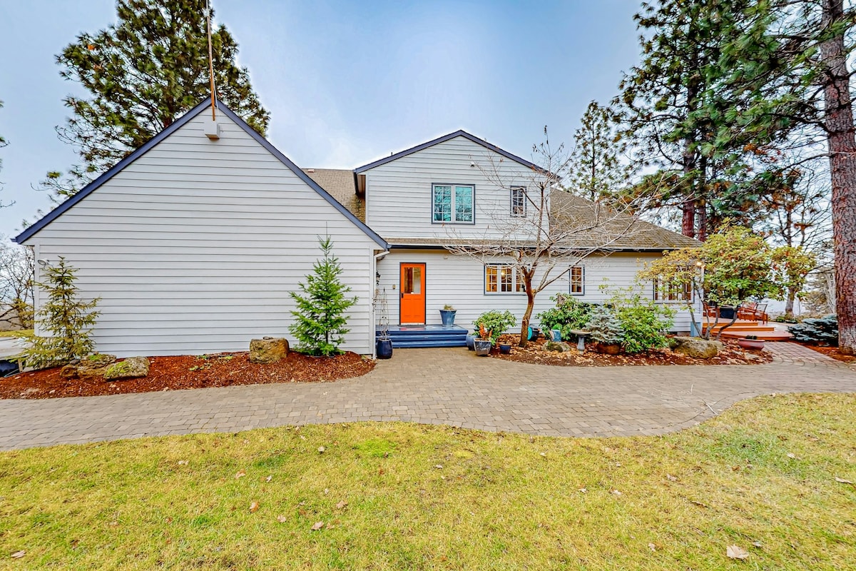 A spacious home is presented with a welcoming orange front door, surrounded by well-maintained landscaping featuring evergreen trees and shrubs. The stone pathway leads to a large patio area, inviting guests to enjoy the outdoor surroundings.