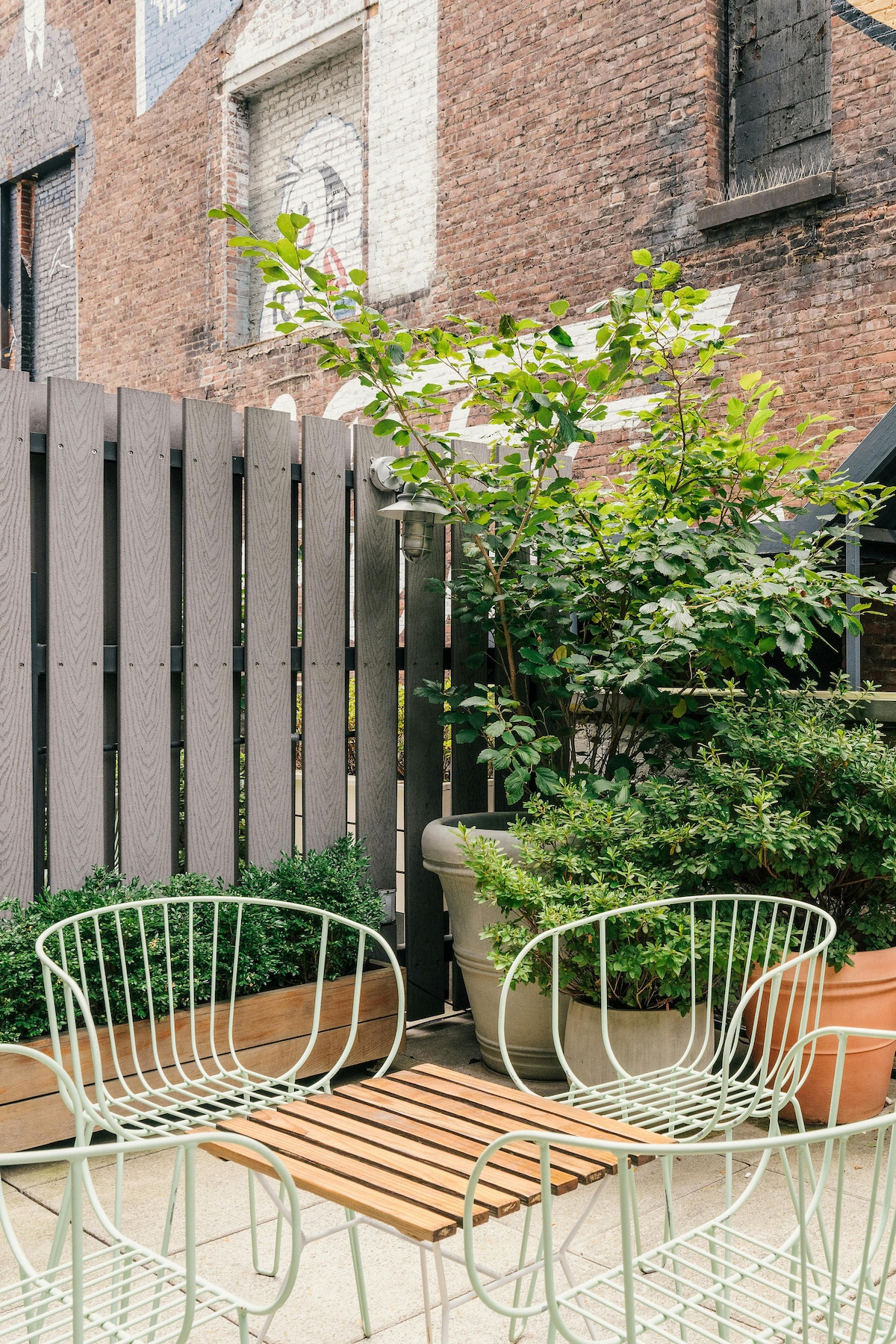 A small outdoor seating area features two light green wire chairs positioned near a wooden slatted table. Lush greenery is present in various pots, with a wooden fence creating a sense of privacy against the brick wall backdrop.