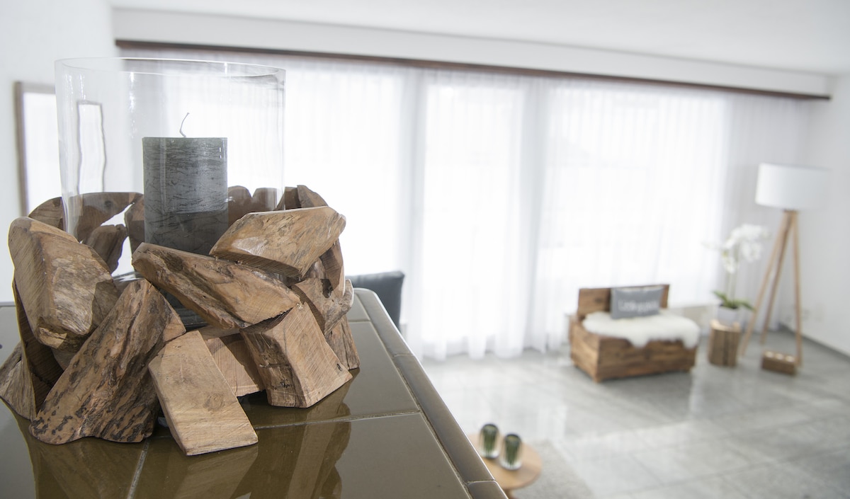 A decorative centerpiece composed of natural wood pieces surrounds a glass candle holder. Soft natural light filters through sheer curtains, illuminating the spacious living area, which includes a side table and a stylish floor lamp.