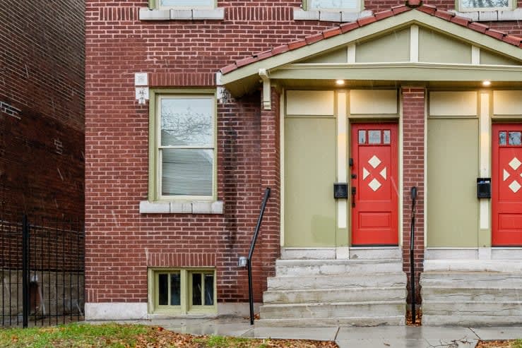 The exterior of a red-brick duplex is shown, featuring two welcoming entrances with bold red doors adorned with diamond-shaped windows. Steps lead up to the doors, and a small grassy area is visible in front, bordered by an iron fence.