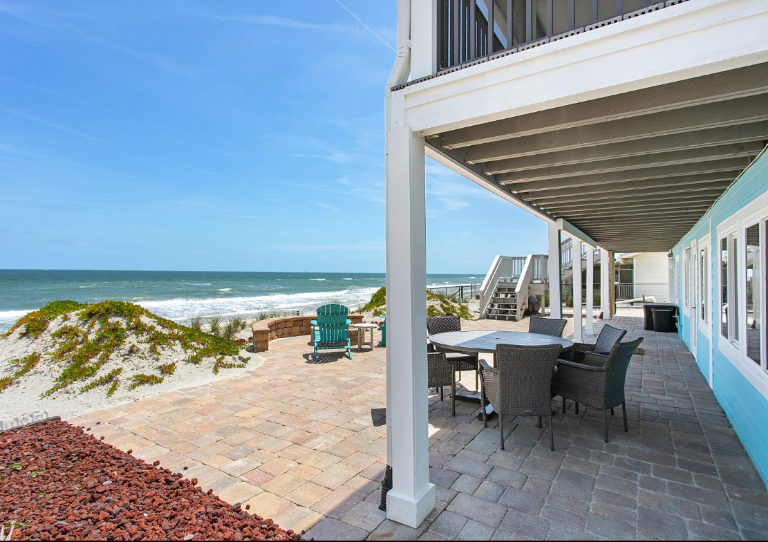 An outdoor patio area is visible, featuring a large dining table surrounded by four chairs. Adirondack chairs are positioned nearby. The beach is just beyond, with gentle waves lapping against the shore. The space is framed by a light blue exterior wall and natural landscaping.