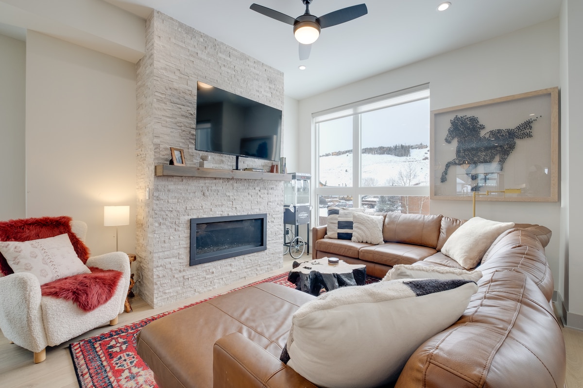 A spacious living area is highlighted by a textured stone fireplace and a large television mounted above it. Soft seating includes a brown leather sectional and a cozy armchair. Natural light fills the room through expansive windows, offering views of the snowy outdoors.