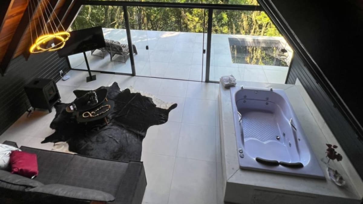 A spacious interior showcases an inviting lounge area with a contemporary couch and a decorative rug. Natural light fills the space through large glass doors, revealing an external deck with a heated jacuzzi nearby. A television stands unobtrusively in the corner, enhancing relaxation.