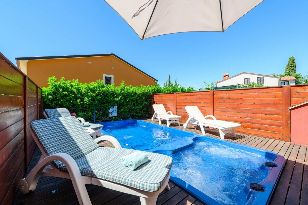 A private courtyard is shown, featuring a small pool equipped with hydromassage jets surrounded by wooden decking. Two loungers are positioned beside the pool, shaded by a large umbrella. Lush greenery forms a natural barrier, ensuring privacy and a peaceful atmosphere.