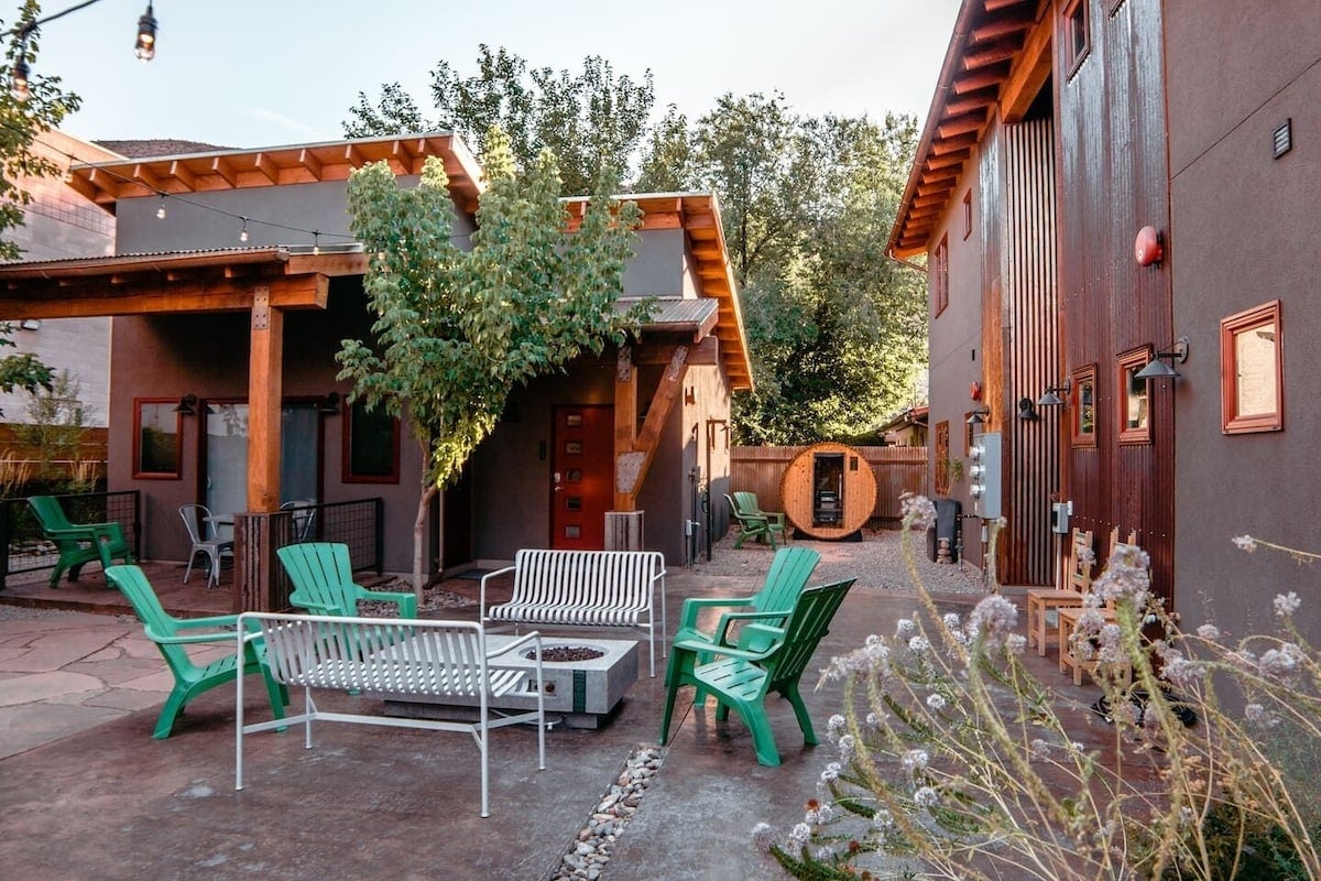 An inviting outdoor space features green lounge chairs and a white seating area, surrounded by trees and rustic buildings. A communal sauna is situated nearby, contributing to a relaxed atmosphere. Decorative plants add a touch of nature to the landscaped surroundings.