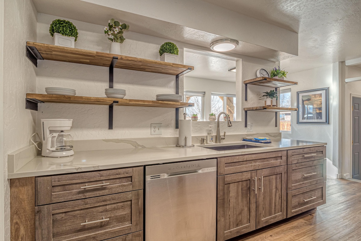A modern kitchen design features a spacious island with a shiny dishwasher and dark wooden cabinets. Open shelving displays decorative plants, while a coffee maker is positioned on the counter. Natural light streams in through nearby windows, brightening the neutral tones.