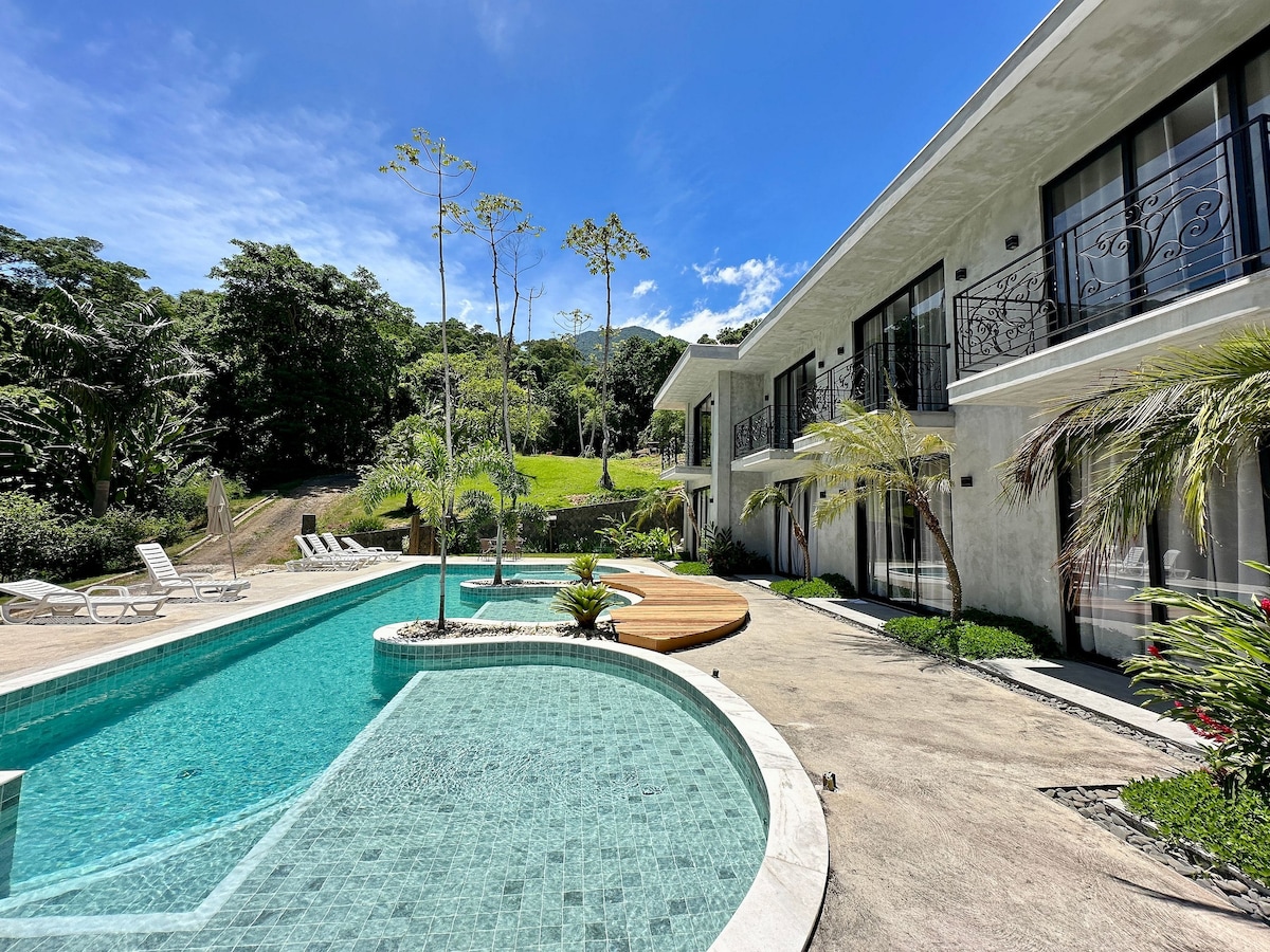 A modern flat is positioned beside a clear swimming pool, surrounded by lush greenery. Sun loungers are placed near the pool's edge, and a wooden deck enhances the outdoor space. Large windows line the facade, offering ample natural light.
