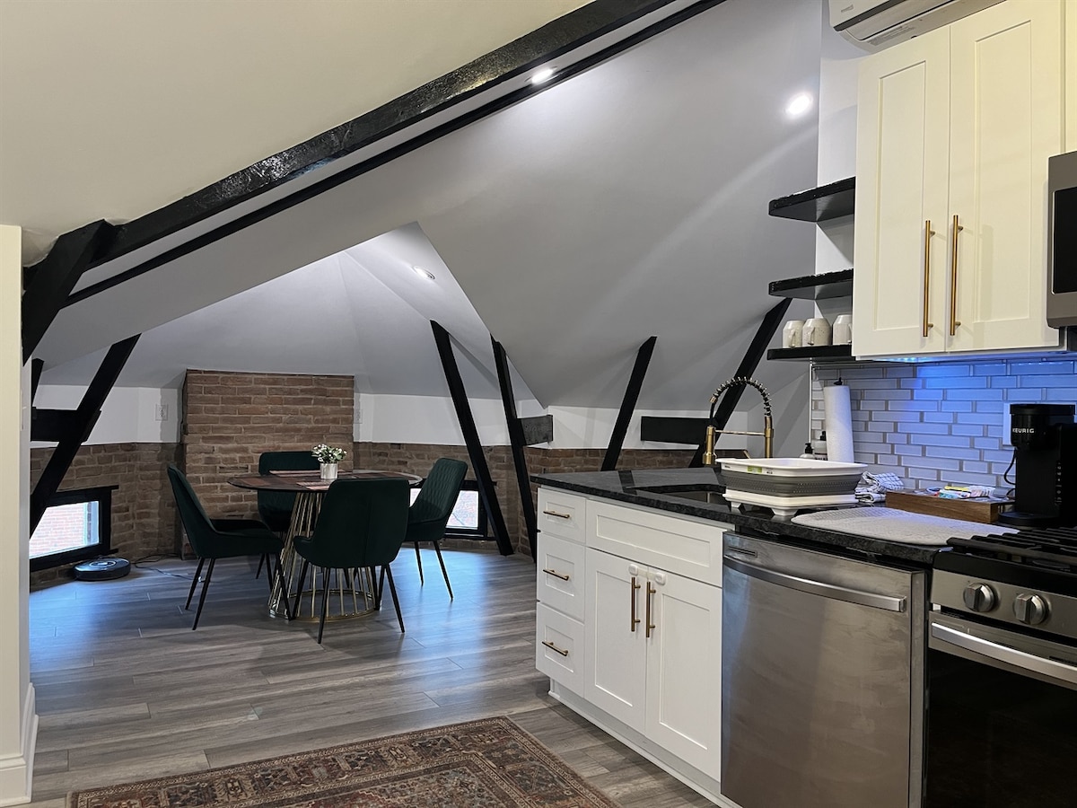 A modern kitchen area is visible, featuring sleek white cabinetry and stainless steel appliances. A cozy dining space with a round table and green upholstered chairs is illuminated by natural light from small windows. Exposed brick walls add texture to the open layout.