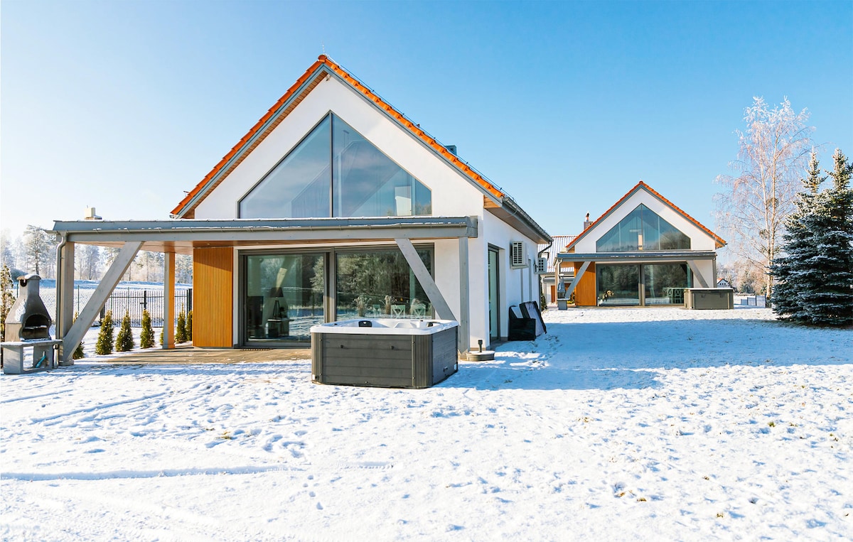 A modern vacation home is presented in a snowy landscape, featuring large glass windows and a wooden deck. A hot tub is positioned outside, surrounded by neatly arranged shrubs. The adjacent building mirrors the architectural style, emphasizing the spacious outdoor area under a clear blue sky.